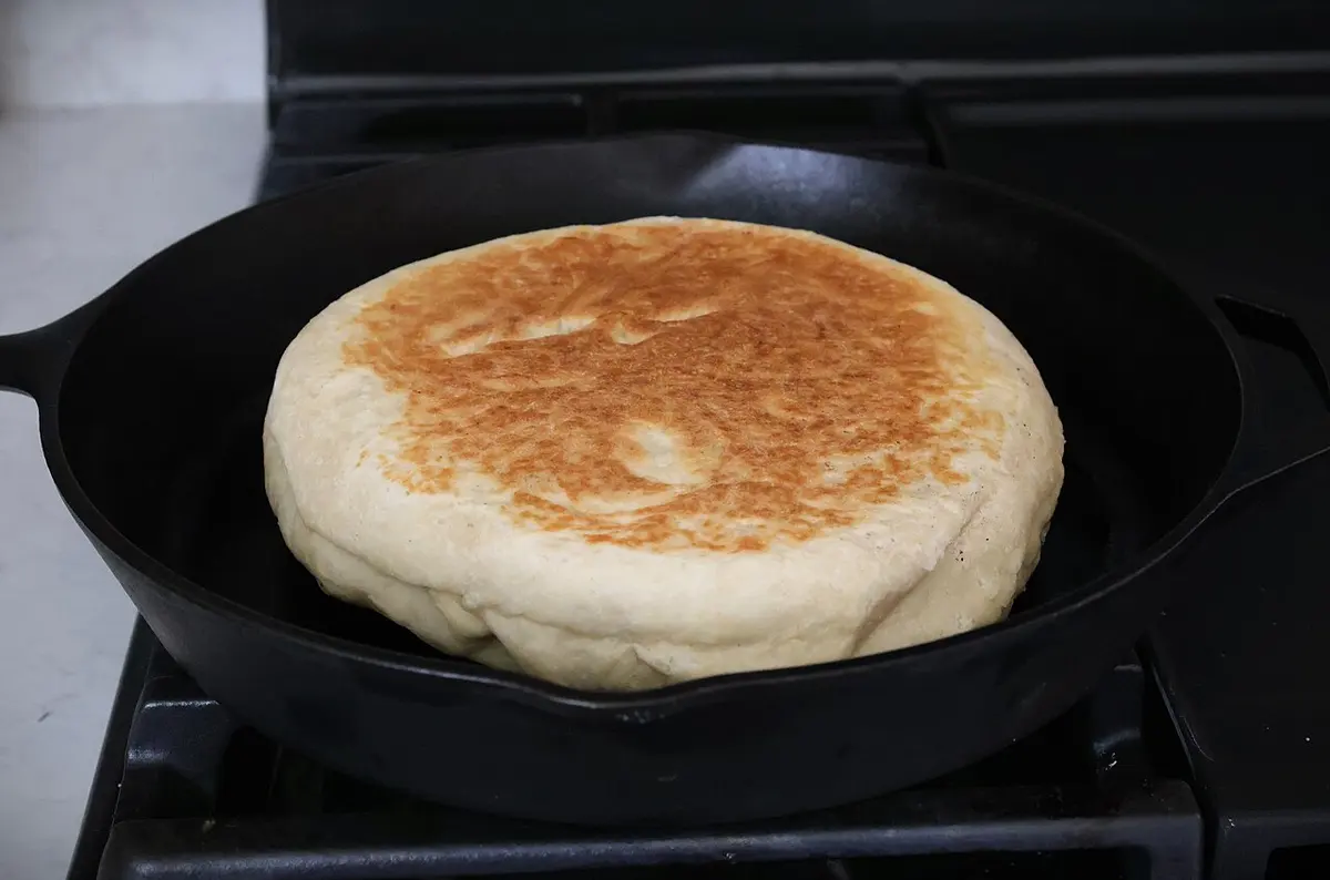 Pão redondo e dourado assando numa frigideira de ferro sobre o fogão