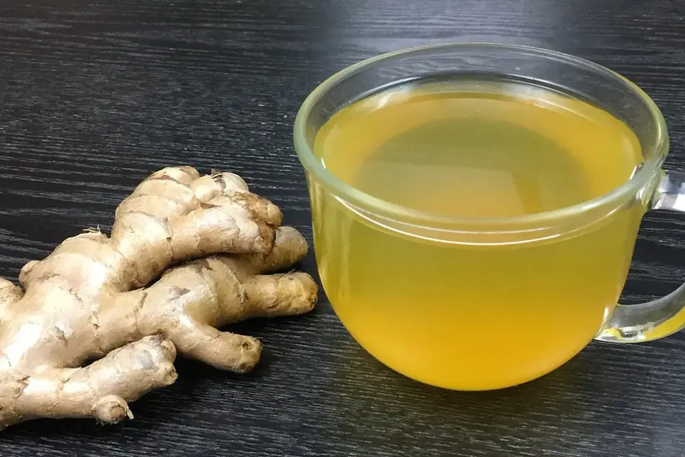 Xícara de vidro com chá de gengibre amarelado ao lado de uma raiz de gengibre fresca